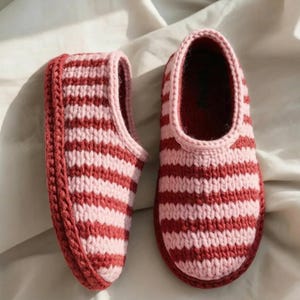 Può includere: Un paio di pantofole a righe fatte a mano. Le pantofole sono lavorate a maglia con fasce alternate di filato rosa chiaro e rosso scuro. Le suole e i bordi sono di un rosso scuro uniforme. Le pantofole sono progettate per l'uso in casa.