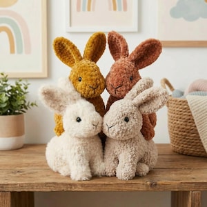 May include: Four plush bunny toys in cream, tan, mustard, and rust colors are arranged on a wooden surface. The bunnies have long ears, black eyes, and stitched facial features. The background includes framed artwork and a woven basket.