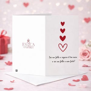 May include: White greeting card with red hearts and Italian text. The card displays a logo and the phrase "Sei un folle a seguire il tuo cuore o sei un folle a non farlo?" on a pink background with heart-shaped confetti.