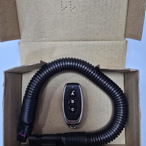Może przedstawiać: Czarny pilot z trzema przyciskami oznaczonymi A, B i C oraz czarna rura falista znajdują się w kartonowym pudełku. Pilot ma srebrną obwódkę. Pudełko jest otwarte, ukazując zawartość.
