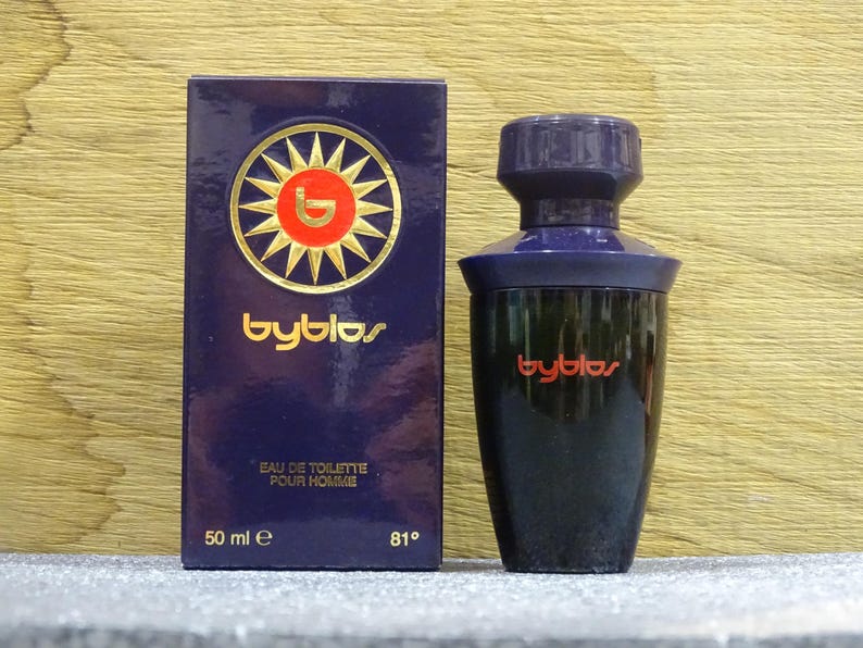 Puede incluir: Un juego de botella y caja de perfume azul oscuro. La caja presenta el nombre de la marca "byblos" en letras doradas, junto con un dise&ntilde;o de sol. La botella es negra con una tapa azul oscuro y el nombre de la marca en rojo.