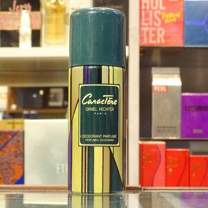 Könnte beinhalten: Eine zylindrische Deodorant-Spraydose mit teal-farbenem Deckel und einem gold-, grün- und lila gestreiften Körper. Auf dem Etikett steht "Caractère Daniel Hechter Paris" und "Deodorant Parfumé Perfumed Deodorant". Im Hintergrund Regale mit verschiedenen Parfümboxen.