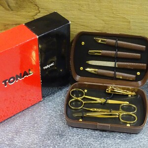 Può includere: Un set per manicure vintage con strumenti dorati e manici marroni, tra cui forbici, tagliaunghie e vari strumenti per la cura della persona. Il set è contenuto in una custodia marrone, accanto a una scatola rossa e nera con la scritta "TONAL" e "TEKNIC".