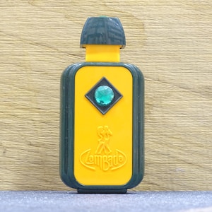 May include: A vintage perfume bottle with a dark green cap and trim, a yellow body, and a green gem. The bottle has the word "Lambada" in a stylized font. The bottle is rectangular with rounded edges.