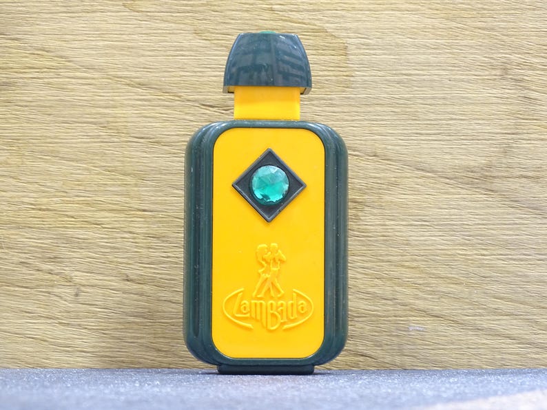 May include: A vintage perfume bottle with a dark green cap and trim, a yellow body, and a green gem. The bottle has the word "Lambada" in a stylized font. The bottle is rectangular with rounded edges.