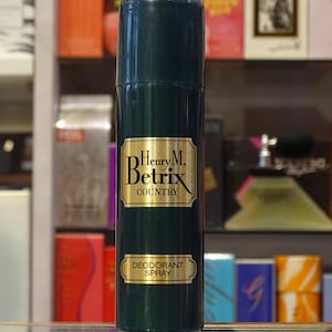 Könnte beinhalten: Eine dunkelgrüne Deodorant-Sprayflasche mit goldfarbenen Buchstaben und Akzenten. Auf dem Etikett steht "Henry M. Betrix COUNTRY DEODORANT SPRAY". Die Flasche ist zylindrisch und steht aufrecht, mit einem Hintergrund aus verschiedenen Parfümflaschen.