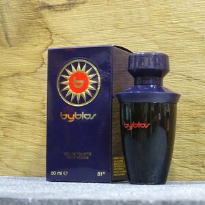 Puede incluir: Un juego de botella y caja de perfume azul oscuro. La caja presenta un dise&ntilde;o de sol dorado con la palabra "byblos" en letras doradas. La botella es negra con una tapa azul oscuro y el logotipo "byblos" en rojo. La caja dice "Eau de Toilette Pour Homme".