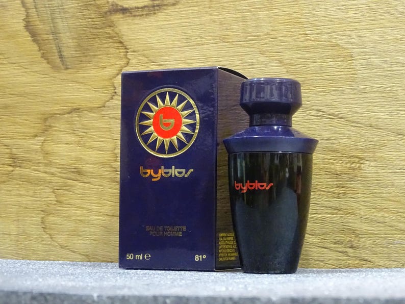 Puede incluir: Un juego de botella y caja de perfume azul oscuro. La caja presenta un dise&ntilde;o de sol dorado con la palabra "byblos" en letras doradas. La botella es negra con una tapa azul oscuro y el logotipo "byblos" en rojo. La caja dice "Eau de Toilette Pour Homme".
