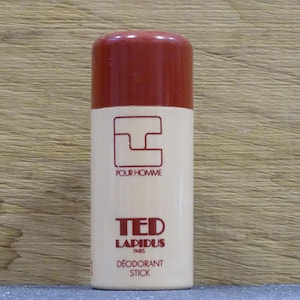 Può includere: Un deodorante in stick cilindrico con tappo bordeaux e corpo color crema. L'etichetta presenta un logo rosso, la scritta "POUR HOMME" e il marchio "TED LAPIDUS PARIS". Il prodotto è etichettato come "DÉODORANT STICK".