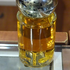 May include: A clear glass perfume bottle filled with amber liquid. The bottle has a gold-colored base with the letters "STC" and a silver and black cap. The bottle is sitting on a reflective surface.