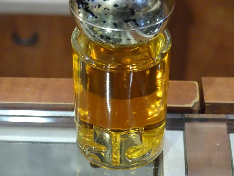 May include: A clear glass perfume bottle filled with amber liquid. The bottle has a gold-colored base with the letters "STC" and a silver and black cap. The bottle is sitting on a reflective surface.