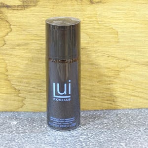 Può includere: Un flacone spray deodorante marrone scuro con la scritta "lui ROCHAS" in bianco. Il flacone è cilindrico e ha un tappo argentato. Il prodotto è un deodorante antitraspirante, peso netto 160 ml.