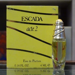 Puede incluir: Un frasco de Eau de Parfum Escada Acte 2 con tapón plateado y caja amarilla. La caja tiene líneas diagonales negras y el texto "Escada acte 2". El frasco contiene 4 ml de perfume.
