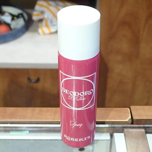 Può includere: Un flacone spray di deodorante rosa con tappo bianco. Il flacone presenta le parole "DEODORO De Luxe" in un design circolare bianco, con "Spray" e "ROBERTS" sotto. Lo sfondo include un mobile in legno e una mensola con vari oggetti.