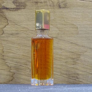 Può includere: Un flacone di profumo in vetro trasparente con un tappo rettangolare color oro. La bottiglia è riempita con un liquido marrone dorato. La bottiglia è su una superficie grigia con uno sfondo di legno.