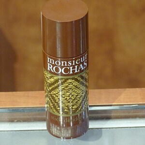 Op de afbeelding: Een bruine deodorantstick met de tekst "monsieur ROCHAS" in het wit. De stick heeft een patroon in goud en bruin. Het product is cilindrisch met een bruine dop.
