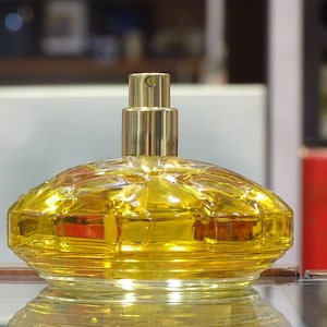 Peut inclure: Un flacon de parfum clair et jaune avec un vaporisateur doré. Le flacon a un design arrondi et texturé et est rempli d'un liquide doré. Le flacon est posé sur une surface réfléchissante.