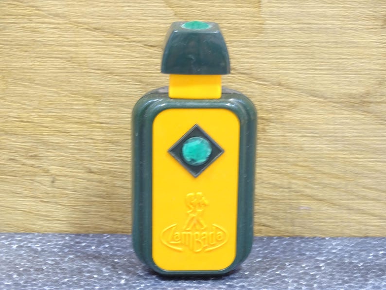 May include: A vintage perfume bottle with a rectangular yellow body and a dark green cap. The bottle features a green gem in a diamond shape and the word "Lombarde" in a curved font.