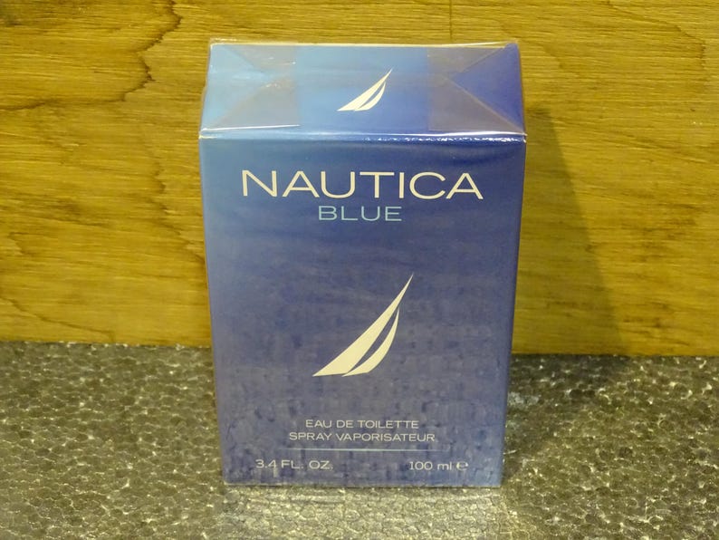 May include: A blue box of Nautica Blue eau de toilette spray. The box features the brand name and logo in white, with the text "EAU DE TOILETTE SPRAY VAPORISATEUR" and "3.4 FL. OZ. 100 ml e" printed on the front.