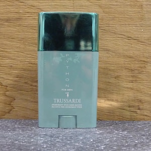 May include: A light teal deodorant stick with a dark teal cap. The product is labelled "PYTHON FOR MEN" and "TRUSSARDI". The bottom of the stick has a small, rectangular base. The background is a wooden surface.