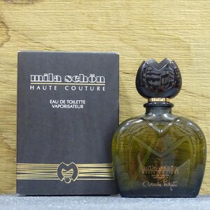 Puede incluir: Un conjunto de perfume con frasco y caja. El frasco es de color verde oliva con un tapón oscuro en forma de flor. La caja es negra con letras doradas que dicen "mila schön HAUTE COUTURE EAU DE TOILETTE VAPORISATEUR". El conjunto está sobre una superficie de madera.