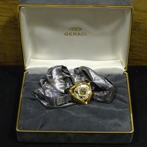 以下が含まれることがあります： 金色のバラのブローチ、銀色の花びら付き。ベルベット張りの箱に入っています。箱はグレーで金色の縁取りがあり、蓋の内側にはブランド名「GERACI」が印刷されています。ブローチが画像の中心です。