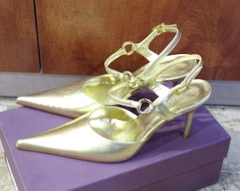 Puede incluir: Un par de zapatos de tacón alto dorados con punta y correas en el tobillo. Los zapatos están sobre una caja morada. Los tacones miden aproximadamente 7,6 cm.