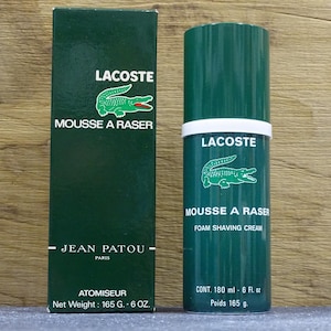 Peut inclure: Une boîte et un flacon de crème à raser Lacoste verts. Le flacon et la boîte présentent le logo Lacoste, un crocodile vert. La boîte indique "MOUSSE A RASER" et "JEAN PATOU PARIS". Le flacon contient 180 ml.