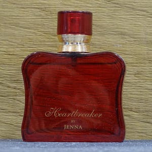 Può includere: Un flacone di profumo rosso con un tappo abbinato e dettagli dorati. Il flacone ha una forma unica e le parole "Heartbreaker by Jenna" sono scritte in oro sul davanti.