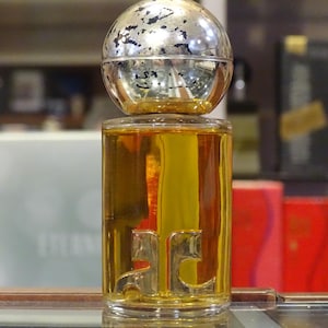 May include: A perfume bottle with a silver and gold spherical cap. The clear glass bottle contains a golden liquid and features a gold emblem. The background includes other perfume boxes.