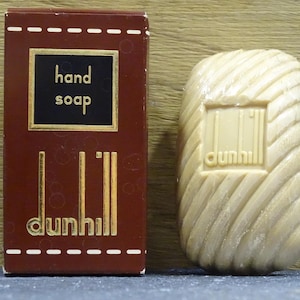 以下が含まれることがあります： 茶色の箱と石鹸。箱には「hand soap」の文字が黒い正方形の中に金色の文字で書かれています。石鹸はベージュ色で、彫刻が施されており、ブランド名は「dunhill」です。