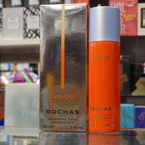 Può includere: Un flacone spray deodorante Rochas Lumiere arancione e la sua scatola abbinata. Il flacone ha un tappo argentato e la scatola ha una finitura argentata con una striscia verticale arancione. Il flacone contiene 100 ml. I testi "Lumiere" e "Rochas" sono stampati.