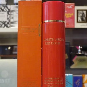 Op de afbeelding: Een rode Elizabeth Arden Red Door deodorant spray en een bijpassende oranje doos. De spuitfles heeft een gouden bandje aan de bovenkant. De doos heeft het Elizabeth Arden-logo en productinformatie. Op de achtergrond staan ​​andere parfumflessen.