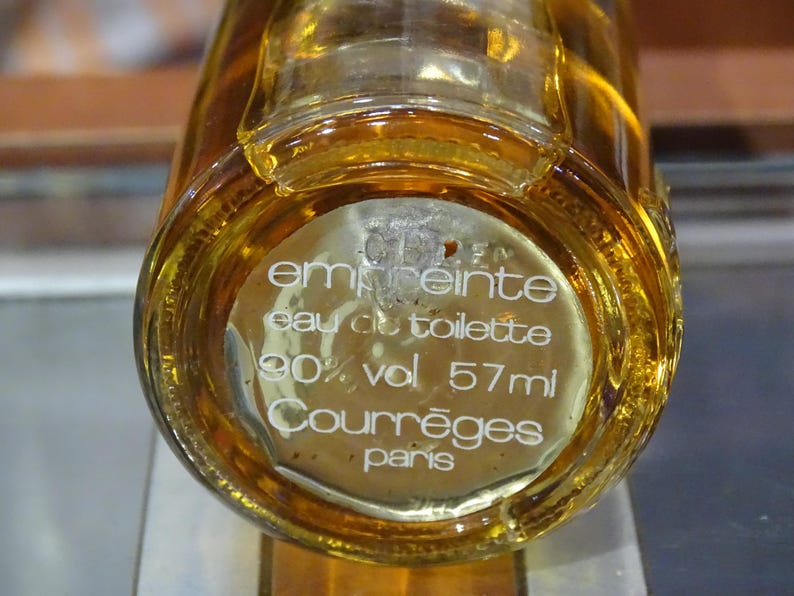 May include: A clear glass bottle of eau de toilette with a golden liquid inside. The label reads "emprunte eau de toilette 90% vol 57ml Courr&egrave;ges paris."