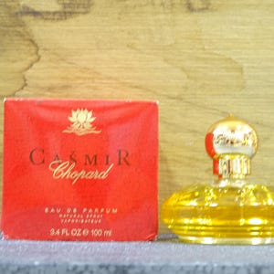 May include: A bottle of Casmir Chopard perfume with a red box. The box has gold lettering and a lotus flower design. The perfume bottle is gold and has a decorative top. The box reads "3.4 FL OZ 100 ml".
