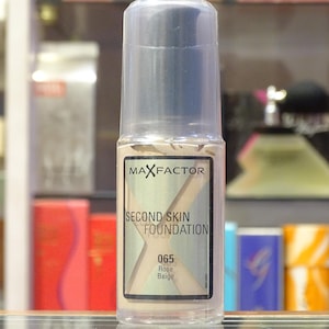 Puede incluir: Un frasco dosificador de plástico transparente de base de maquillaje Max Factor Second Skin en el tono 065 Rose Beige. La etiqueta presenta una letra X grande sobre el texto, con el nombre de la marca arriba y el tono y número debajo.