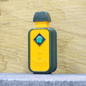 May include: A rectangular, vintage perfume bottle with a yellow front and dark green sides and top. The front features a diamond-shaped accent with a green gem and the word "Lombarde". The bottle is on a gray surface.