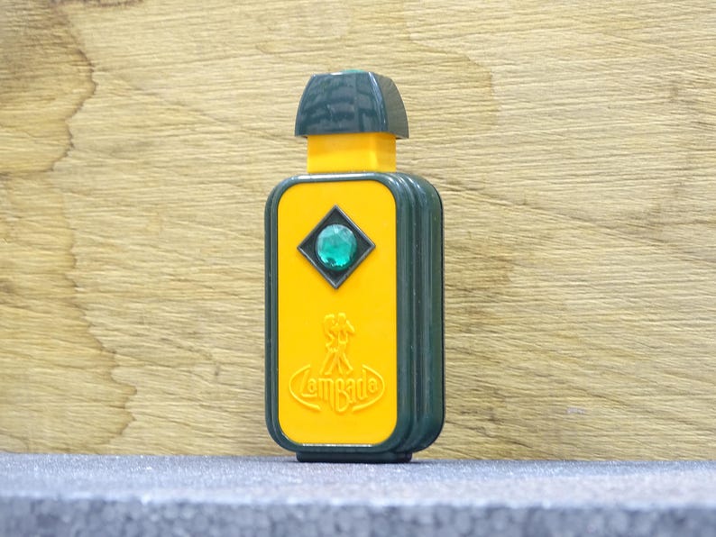 May include: A rectangular, vintage perfume bottle with a yellow front and dark green sides and top. The front features a diamond-shaped accent with a green gem and the word "Lombarde". The bottle is on a gray surface.