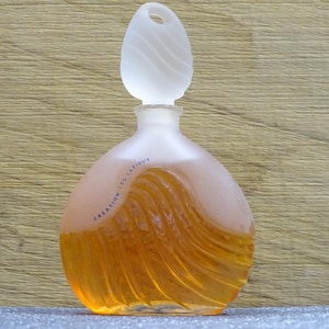 Könnte beinhalten: Ein parfümflakon aus mattiertem Glas mit einem einzigartigen Design. Die Flasche hat eine abgerundete Form mit einem wellenartigen Muster und einem mattierten, tropfenförmigen Verschluss. Die bernsteinfarbene Flüssigkeit ist sichtbar, mit dem Text "CREATION TED LAPIDUS".