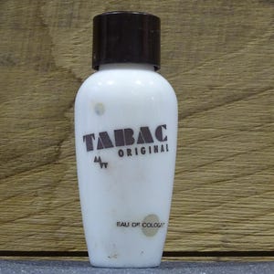 May include: A white Tabac Original Eau de Cologne bottle with a dark brown cap. The bottle features brown text with the brand name and product details. The background is a light brown wooden surface.