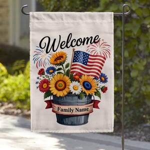 May include: A white garden flag with the word "Welcome" in script. The flag features a floral arrangement in a metal bucket with an American flag and fireworks. A red banner reads "Family Name".