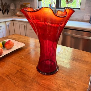 May include: A tall, vibrant red glass vase with a flared, ruffled rim. The vase has a wide opening and a slender, elongated body. It sits on a wooden table, showcasing its elegant design and rich color.