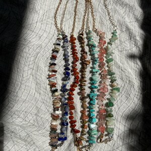 May include: Six bracelets featuring irregular, tumbled gemstone chips in various colors including blue, red, brown, turquoise, pink, and green. Each bracelet is strung on a thin gold-tone metal chain with a lobster claw clasp at the end.