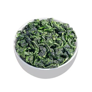 May include: A white ceramic bowl filled with green tea leaves. The tea leaves have a dark green color with lighter green highlights, and are tightly curled. The bowl is set against a white background.