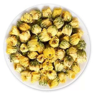 Op de afbeelding: Een witte schaal gevuld met gedroogde chrysantenbloemen. De bloemen zijn een mix van geel en groen, met dicht opeengepakte bloemblaadjes. De afbeelding is een bovenaanzicht, dat de textuur en kleur van de gedroogde bloemen laat zien.