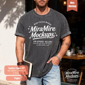 Op de afbeelding: Een donkergrijs t-shirt met opgerolde mouwen met de tekst "MiraMire Mockups" en andere tekst. Het shirt is een mockup voor kledingverkopers. De afbeelding bevat ook een boek en een label "Men Mockup".