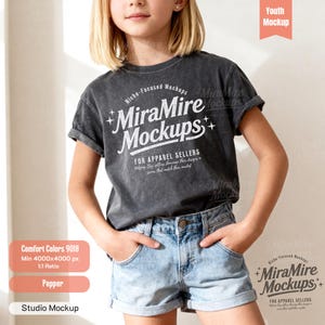 May include: A charcoal gray short-sleeve t-shirt with a vintage-style wash. The front features white text that reads MiraMire Mockups, Niche-Focused Mockups, and For Apparel Sellers, accompanied by small star icons. The shirt is paired with light blue denim shorts.