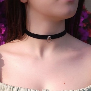Może przedstawiać: Czarny aksamitny naszyjnik typu choker z cienką, gładką opaską, która ściśle przylega do szyi. Środek chokera ozdobiony jest małym, okrągłym, srebrnym metalowym pierścieniem przymocowanym do pasującego srebrnego metalowego łącznika.