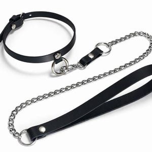 Op de afbeelding: Een zwarte leren halsband met een zilverkleurige metalen ring, verbonden via een zilveren metalen ketting aan een bijpassende zwarte leren riem met een lusgreep. De halsband is voorzien van een verstelbare gespsluiting met zilverkleurig beslag.