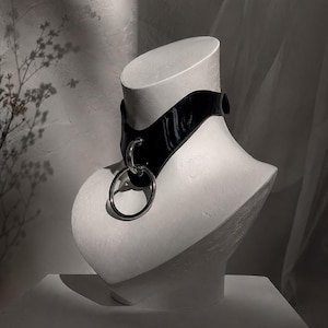 Może przedstawiać: Czarny, błyszczący choker z szerokim paskiem i dużym, srebrzystym metalowym kółkiem przymocowanym do mniejszego okrągłego łącznika z przodu. Obroża jest zaprezentowana na białym popiersiu manekina, aby ukazać jej kształt i konstrukcję.